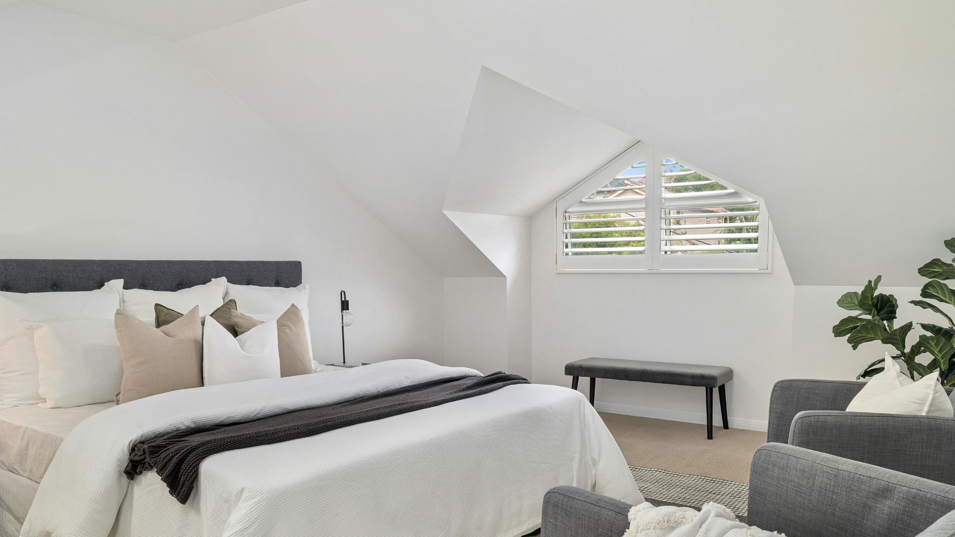 A bright, minimalist bedroom with a large bed, gray accent chairs, a small bench, and a unique arched window.