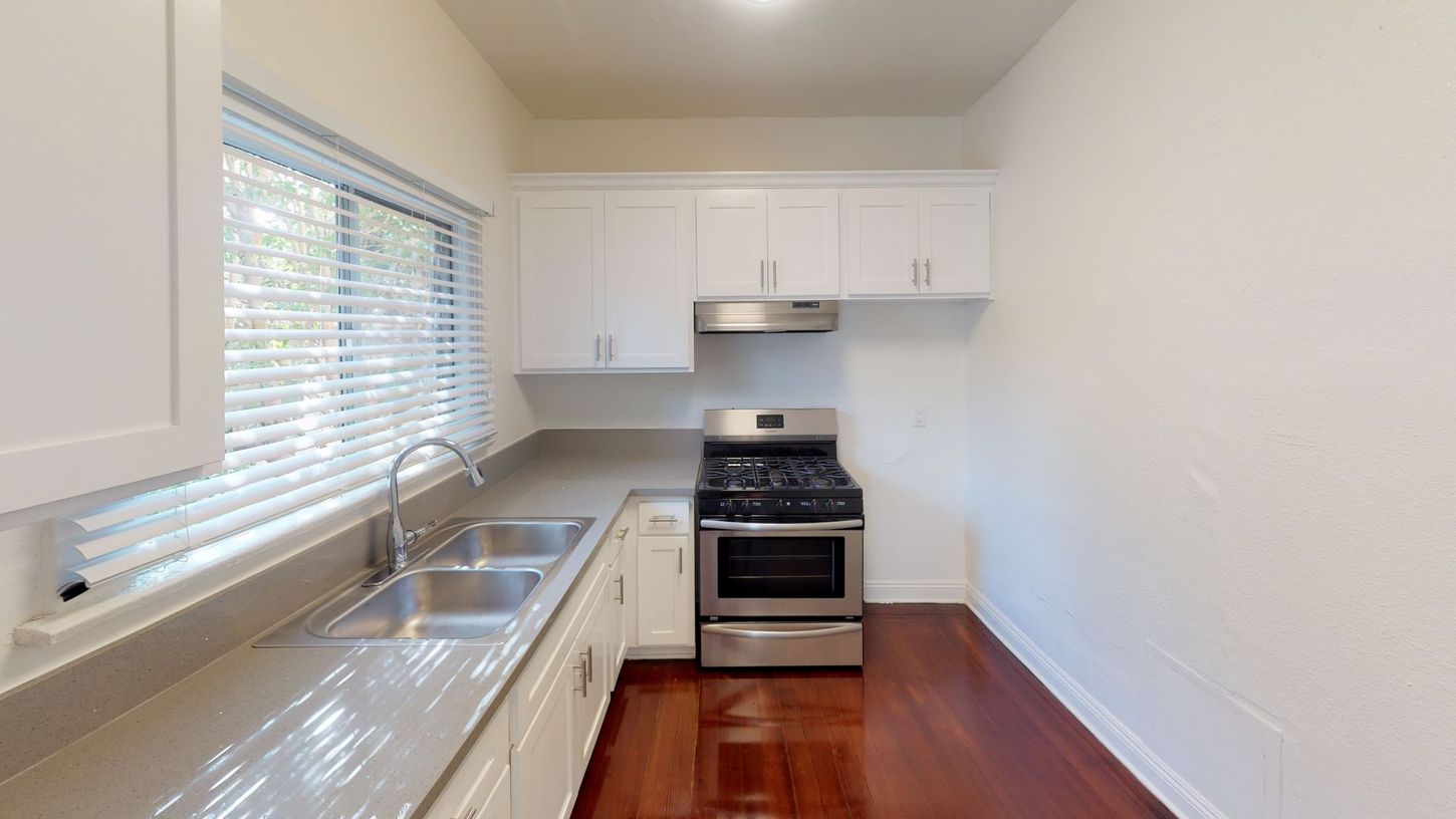 Small, white kitchen with stainless steel appliances, sink by the window, and wood floor.