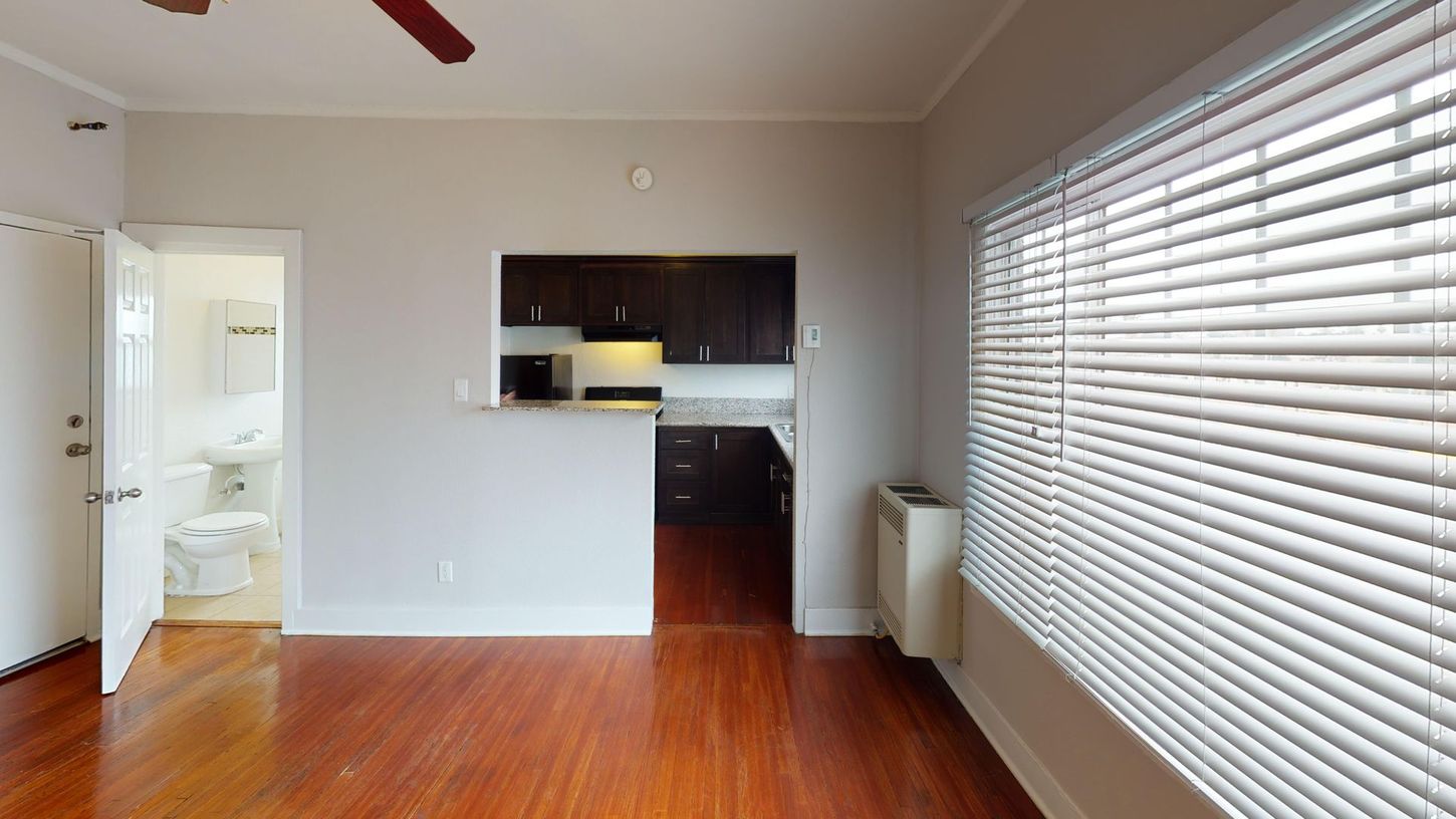Interior of an apartment with hardwood floors, kitchen, open bathroom, and a window with blinds.