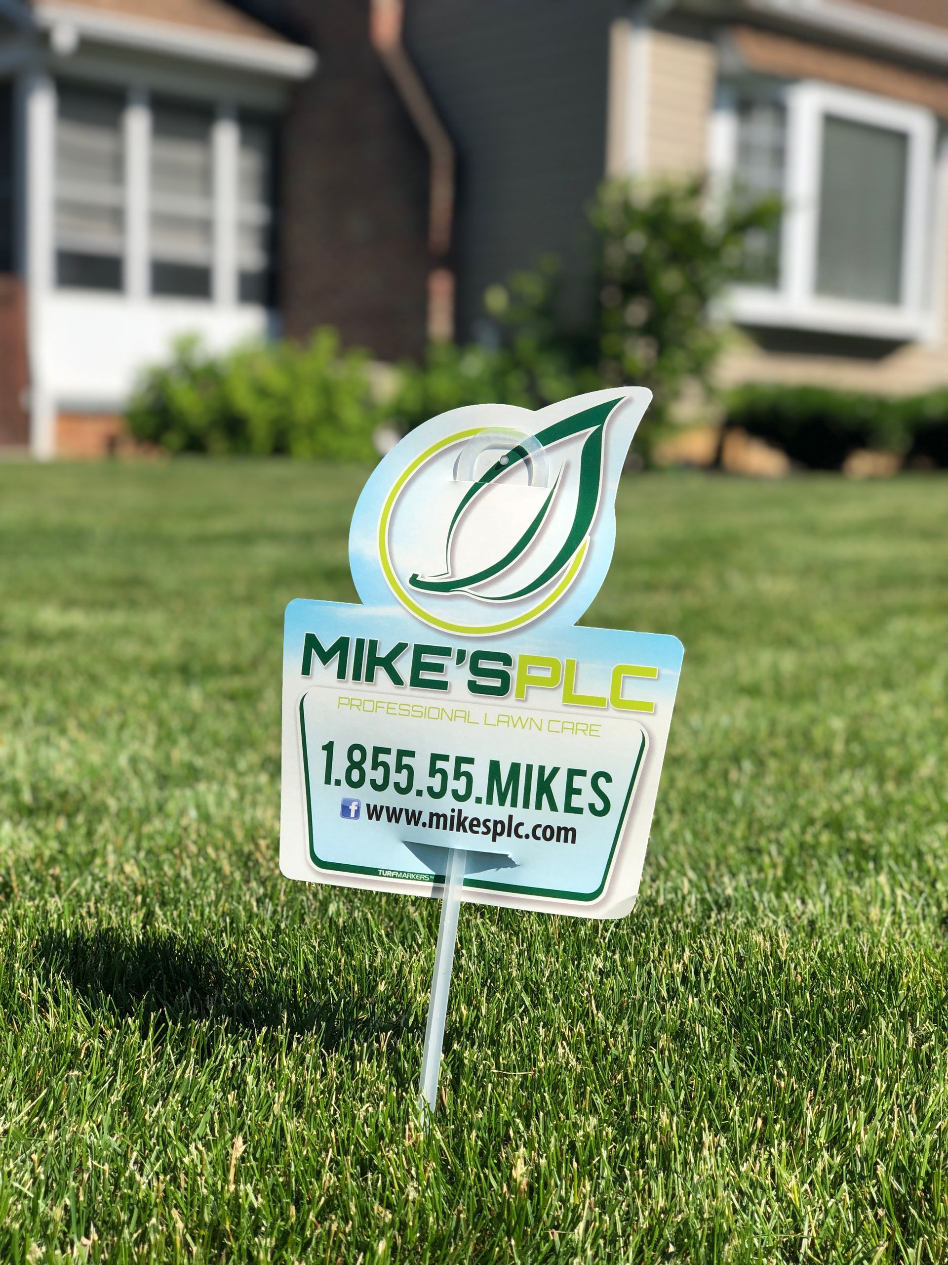 A sign that says `` mike 's plc '' is sitting in the grass in front of a house.