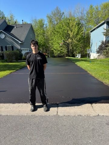 A young man is standing on the side of a paved driveway in front of a house.