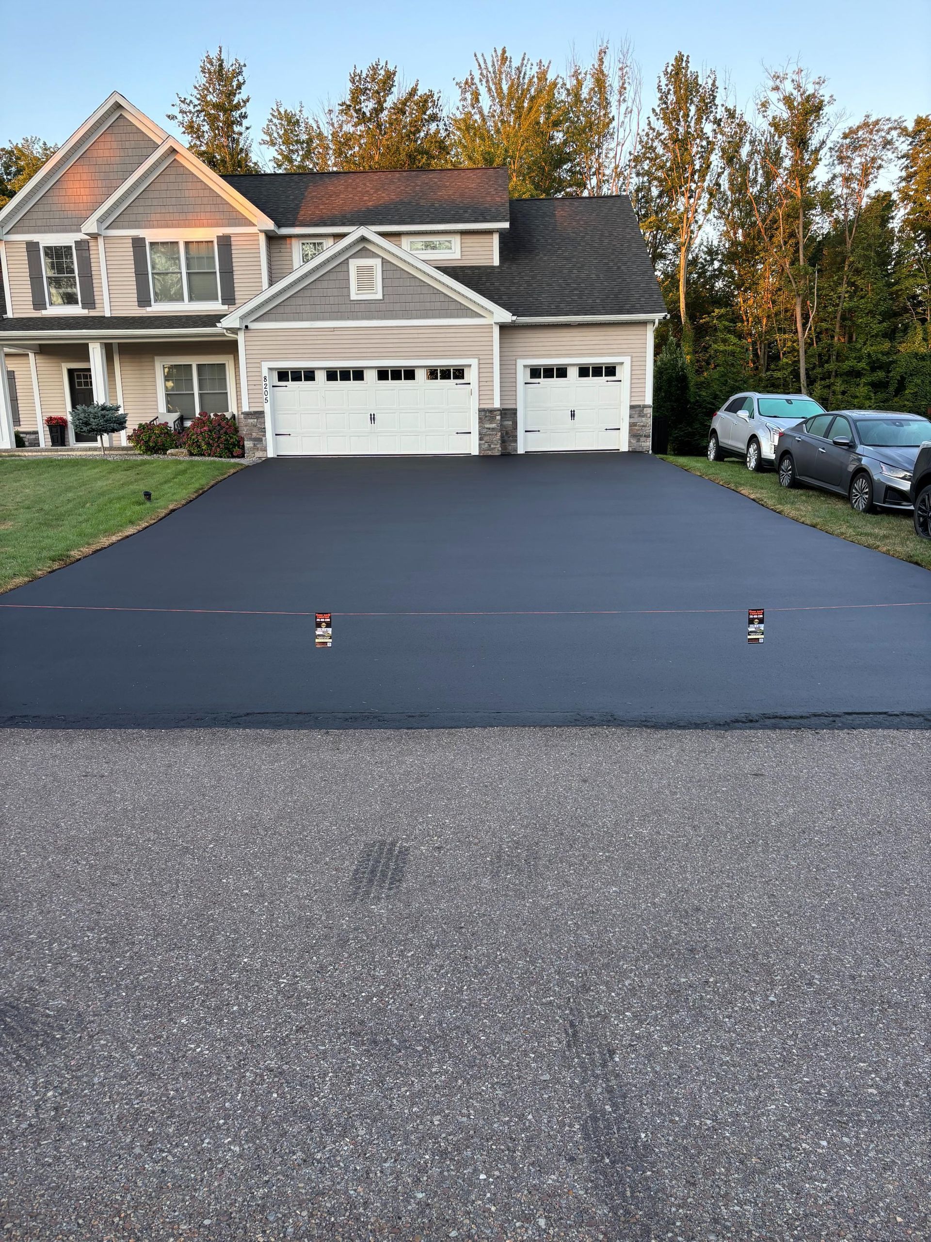 Newly paved black asphalt driveway in front of a two-story house with cars parked on the right.