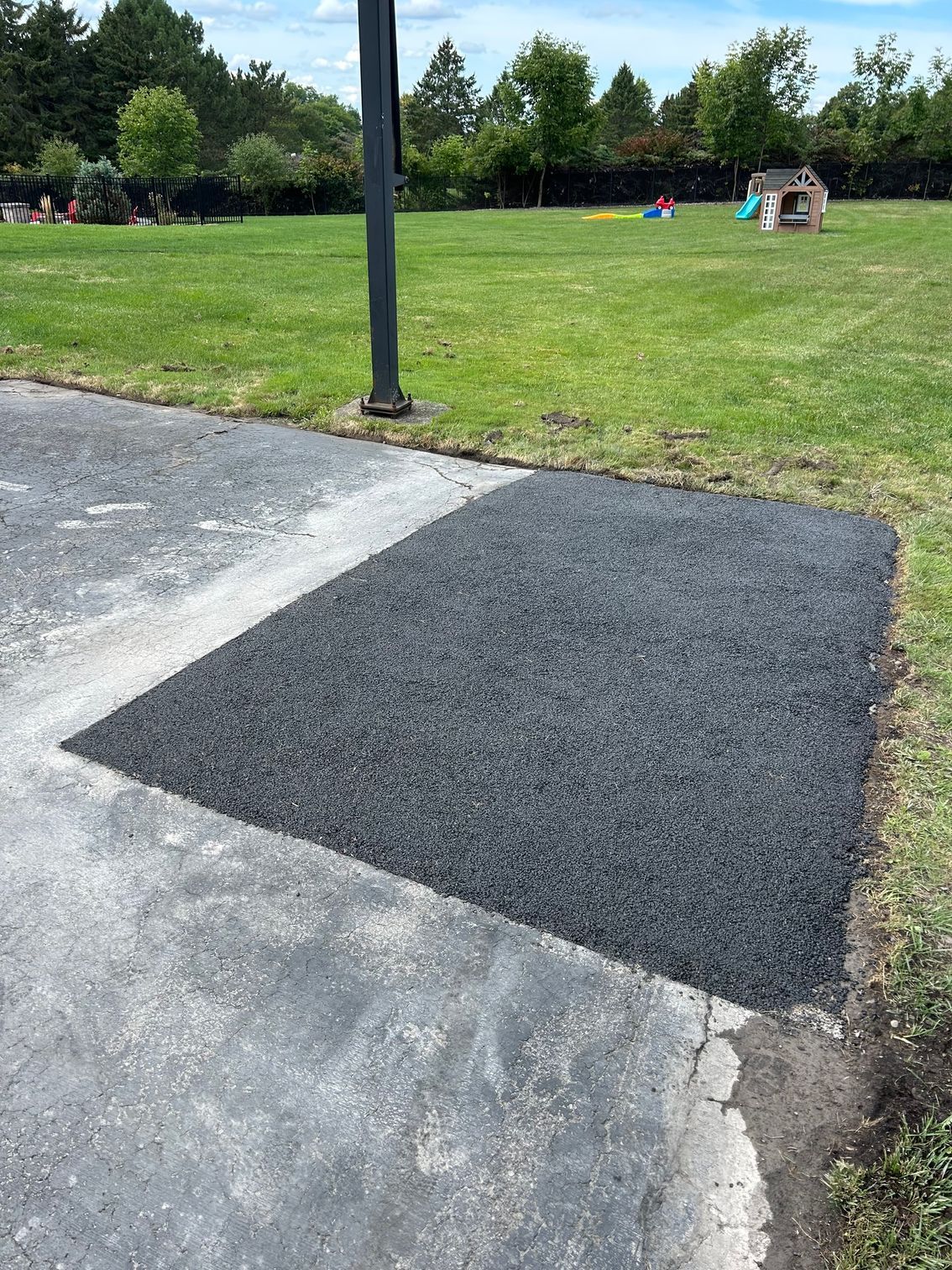 Black rubberized surfacing patch on asphalt, next to grass; basketball hoop in background.