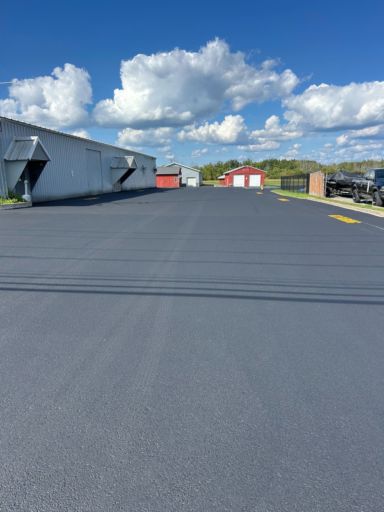 Black asphalt parking lot with gray building on the left and red buildings in the distance under a blue sky.