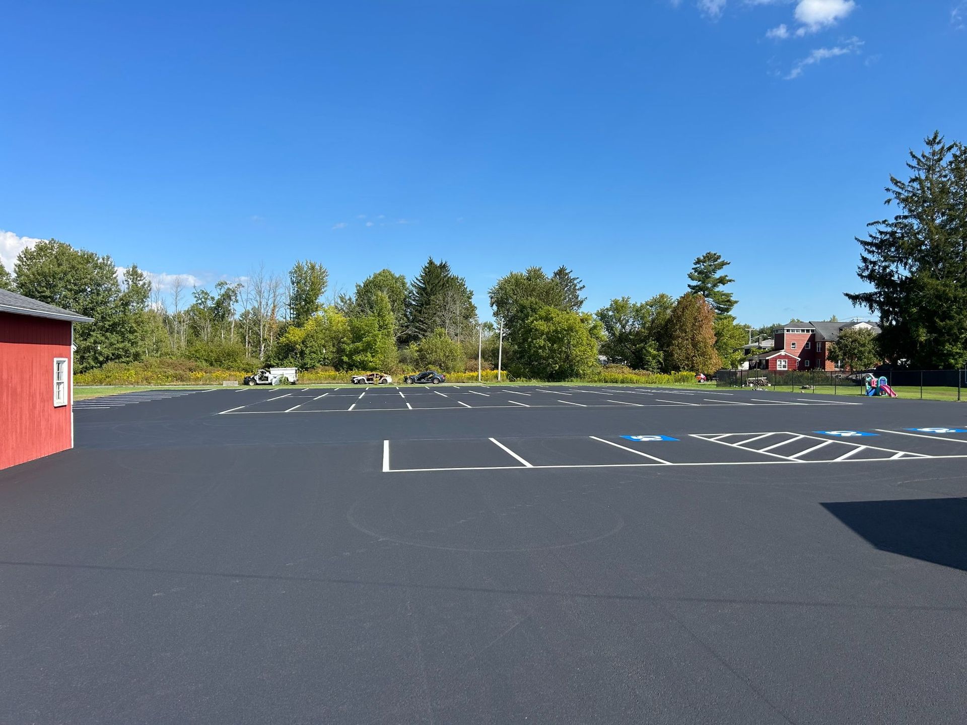 Empty asphalt parking lot with white painted lines, green trees, blue sky, and a red building.