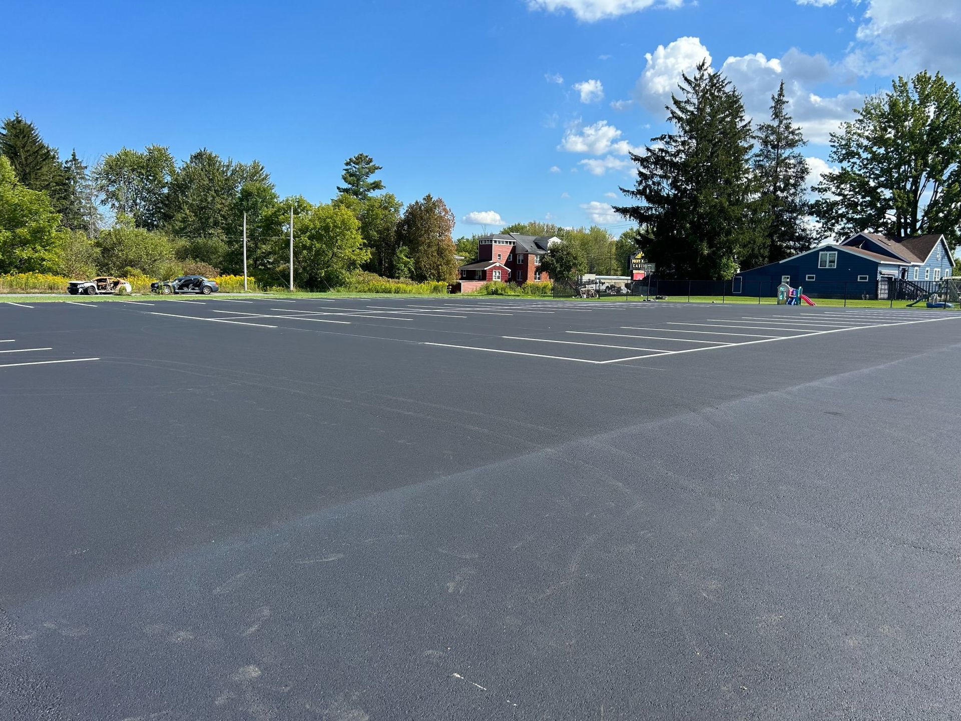 Newly paved black asphalt parking lot with trees and houses in the background under a blue sky with clouds.