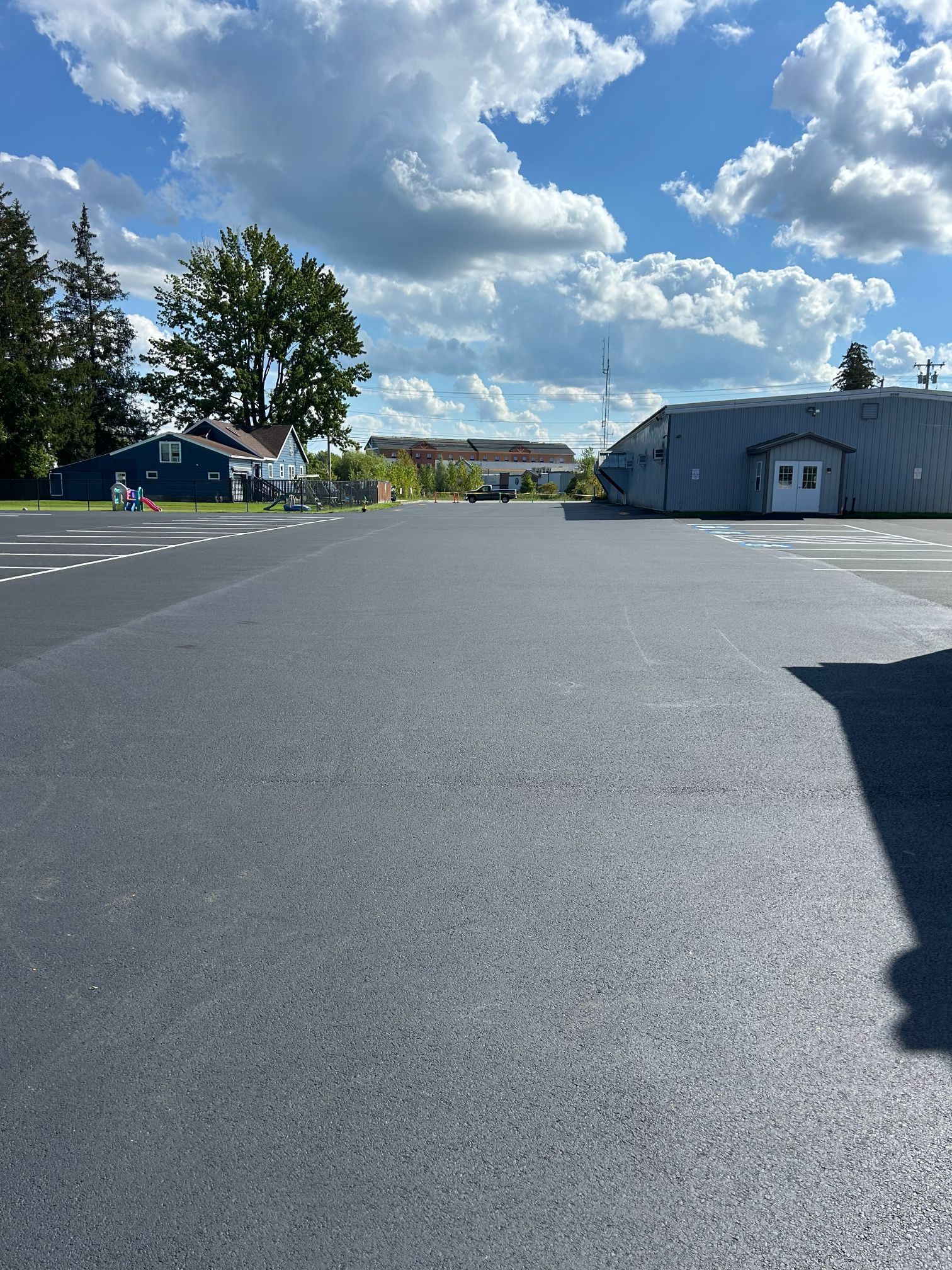 Newly paved parking lot under a blue sky with fluffy clouds, buildings in the background.