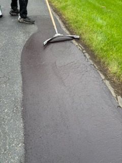 A person is using a broom to spread asphalt on the side of a road.