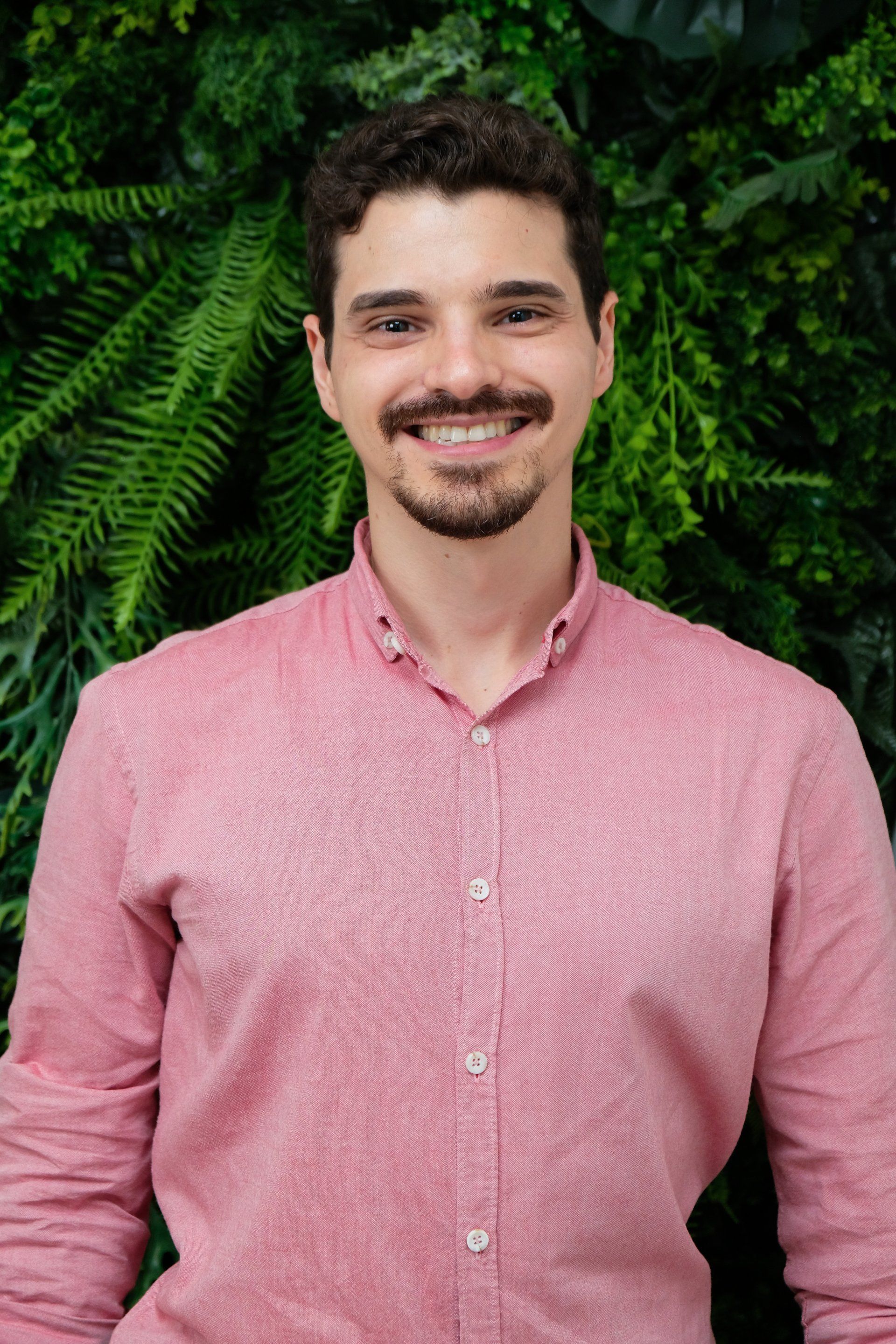 Um homem de camisa rosa está sorrindo em frente a uma parede verde.