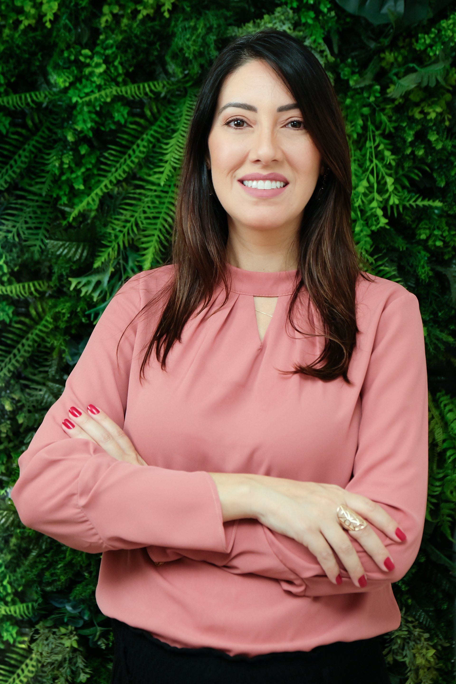 Uma mulher de camisa rosa está parada com os braços cruzados em frente a uma parede verde.