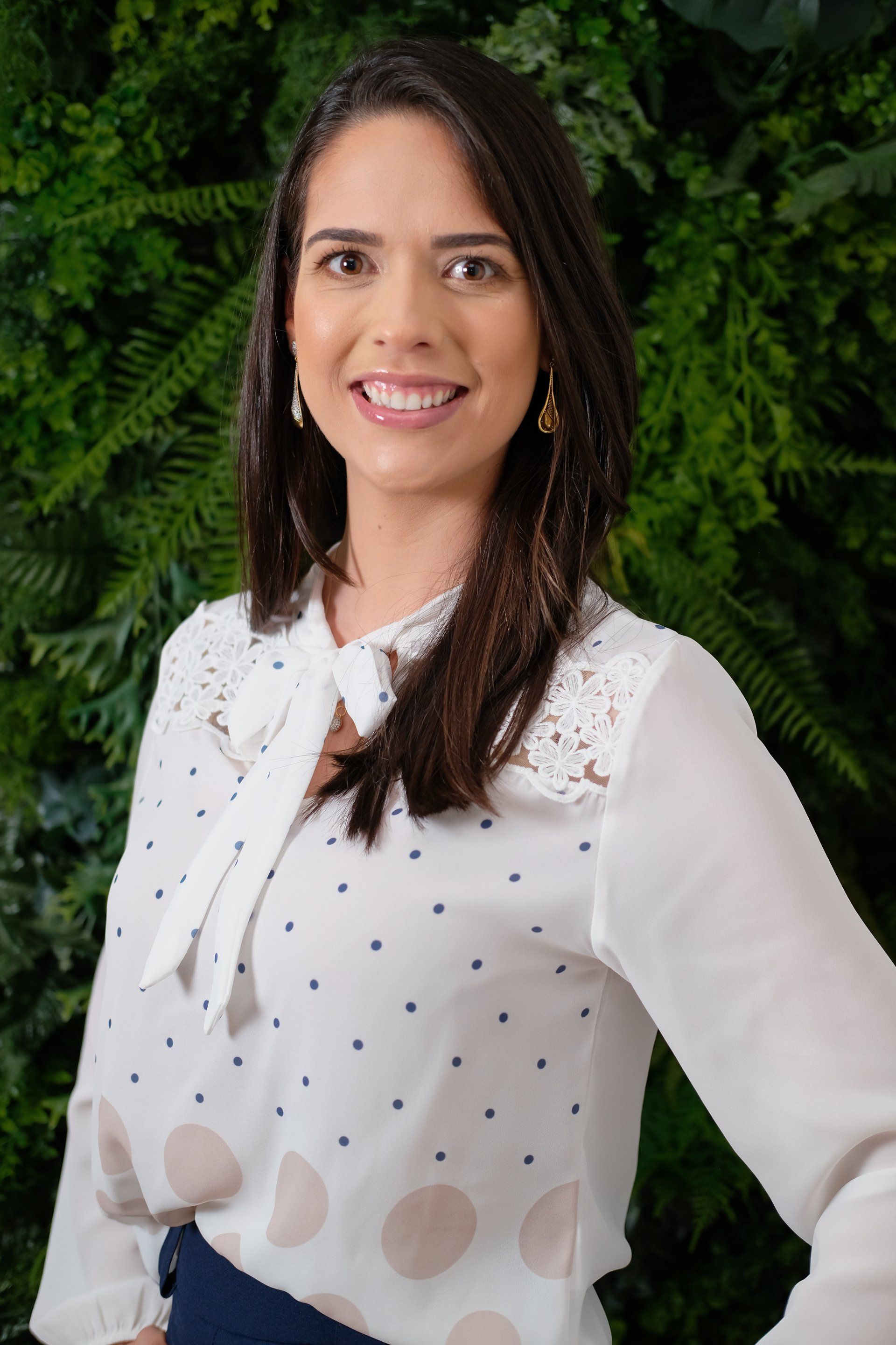 Uma mulher com uma blusa branca de bolinhas está sorrindo em frente a uma parede verde.