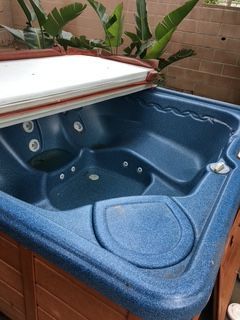 Blue hot tub with partly open white cover, set in a wood frame, against a backdrop of green plants.