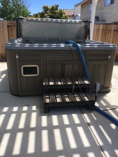 Hot tub outside with open cover, blue hose, and wooden steps on a concrete patio.
