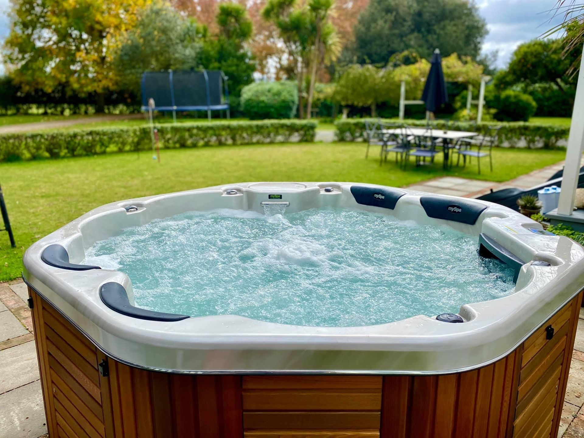 Hot tub with bubbling water on a brick patio in a backyard with green lawn, a table, and trees.