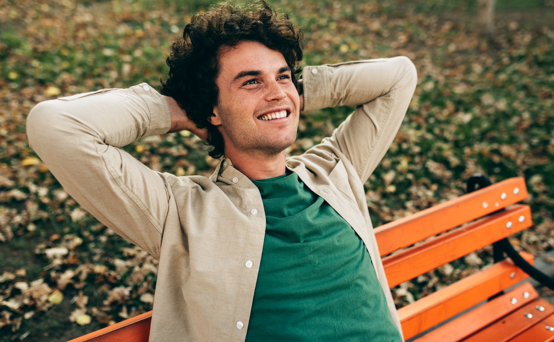 Man smiling with arms behind head, sitting on a park bench, looking up.