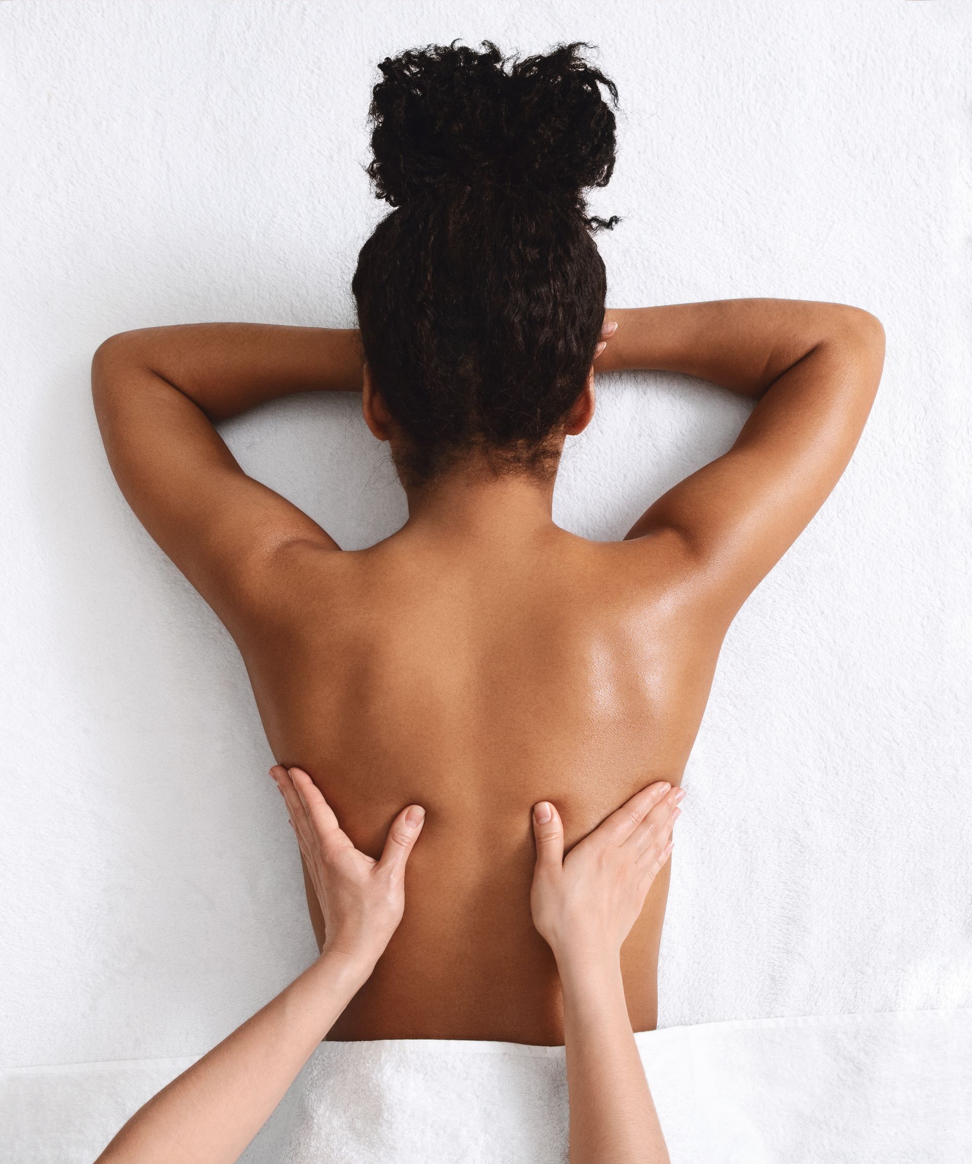 Person receiving a back massage on a white surface. Hands kneading back. Hair in a bun.