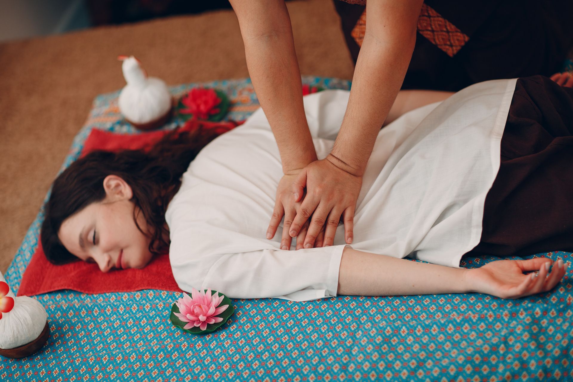 Massage therapist giving a back massage to a person lying on a patterned mat with candles and flowers nearby