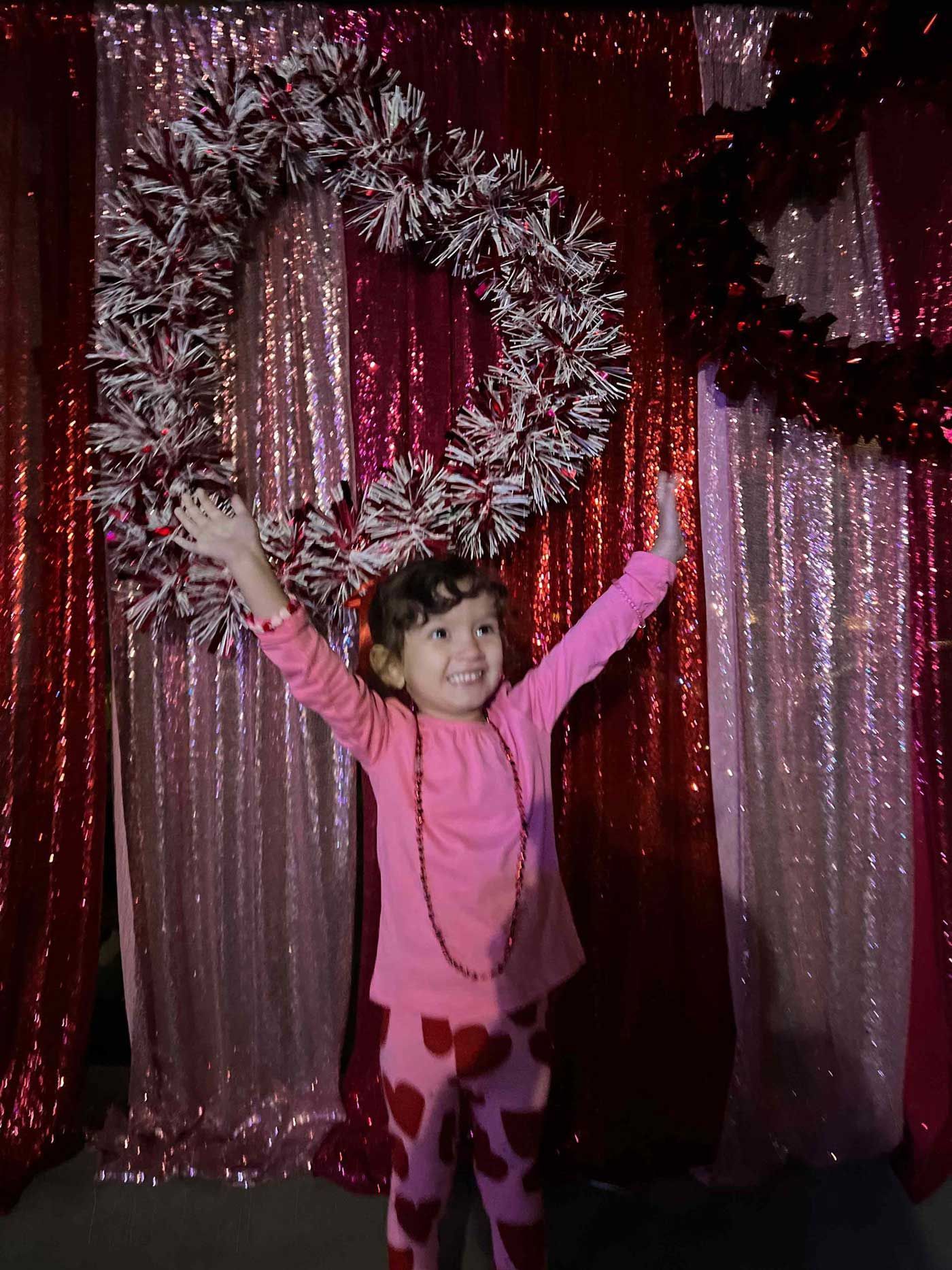 A little girl in a pink shirt is standing in front of a heart shaped wreath.