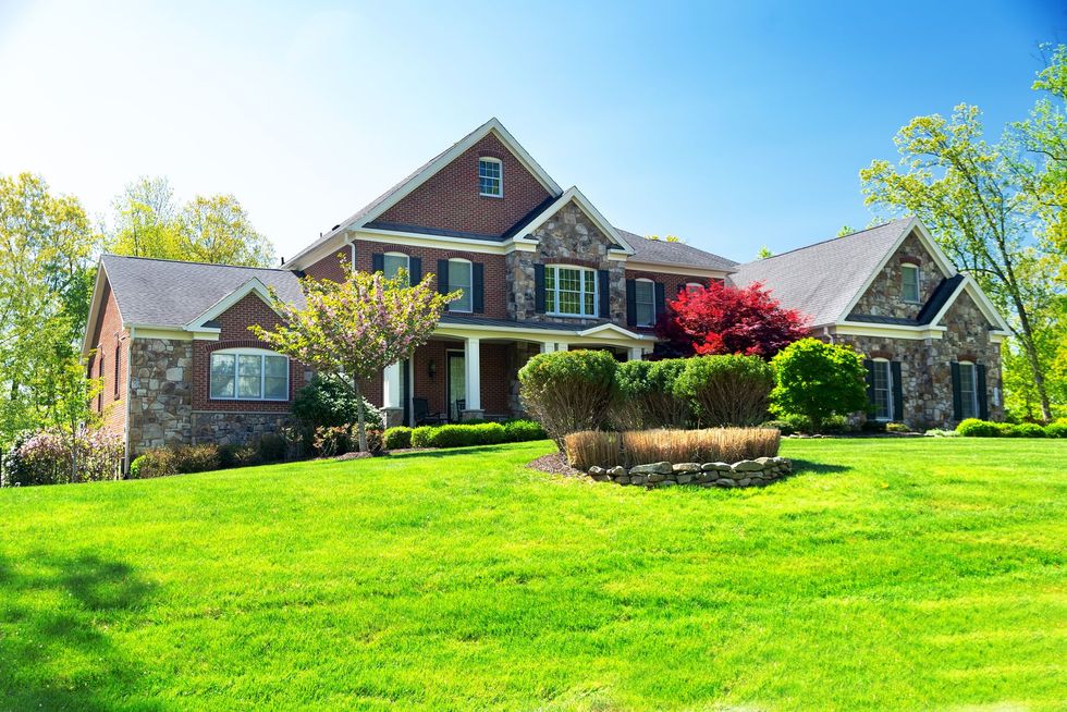 A large, two-story stone and brick house with a dark roof and a green lawn under a clear blue sky.