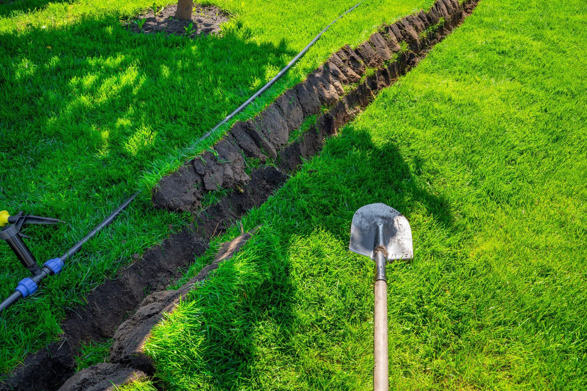 A narrow trench dug in a green lawn with a shovel standing nearby and a pipe running along the grass.