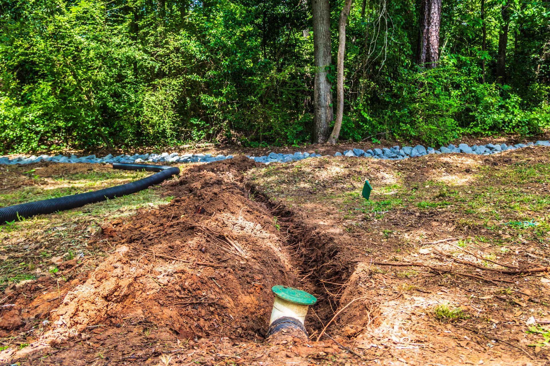 A trench is dug in a yard with a green-capped pipe sticking out of the ground near a wooded area.