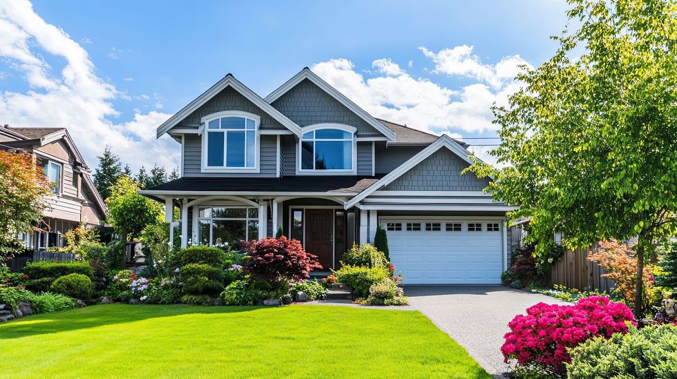 A two-story grey suburban house with a white garage, front lawn, and blooming garden under a clear blue sky.