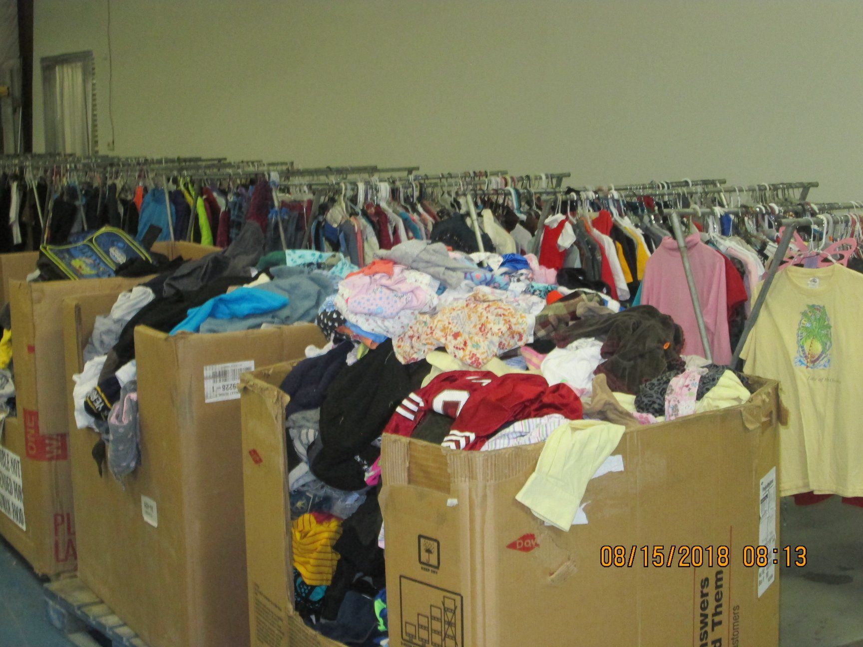 Cardboard boxes overflowing with clothing, clothing racks in background. Warehouse setting.
