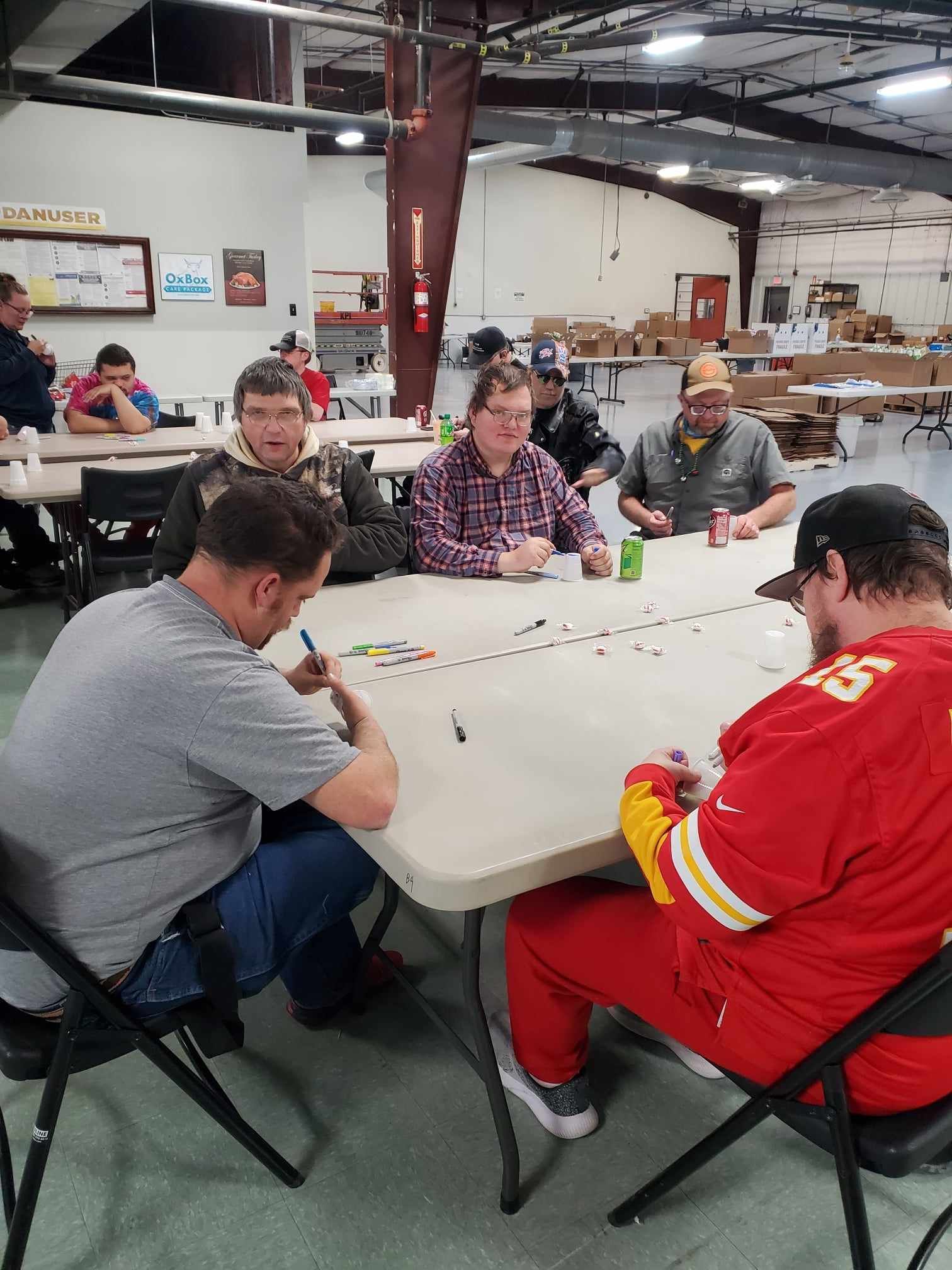 People sitting around a table, playing a game in an indoor setting. Some are writing, with drinks nearby.