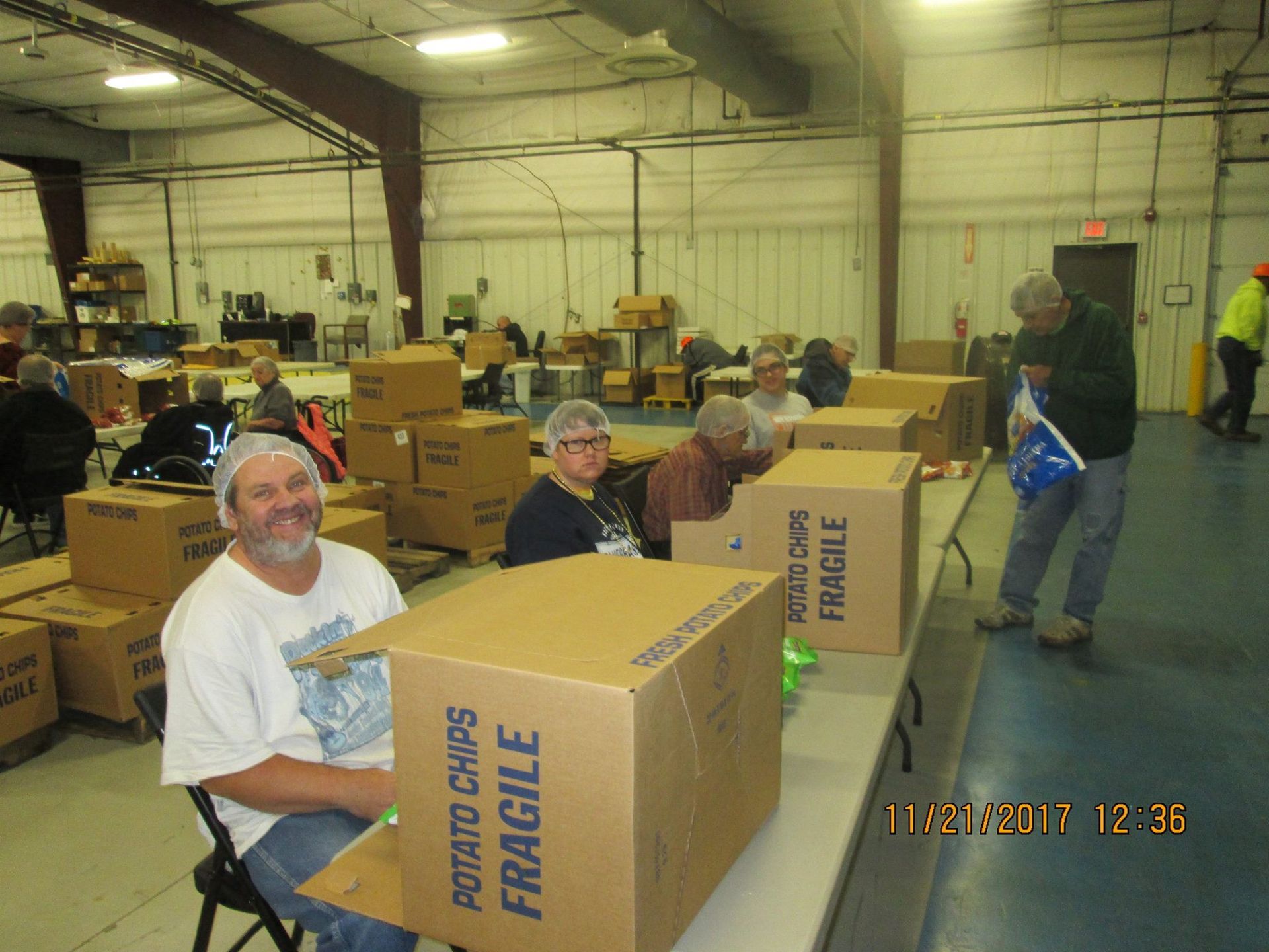 Volunteers packing boxes of potato chips in a warehouse. Several people are seated at tables, smiling and working.