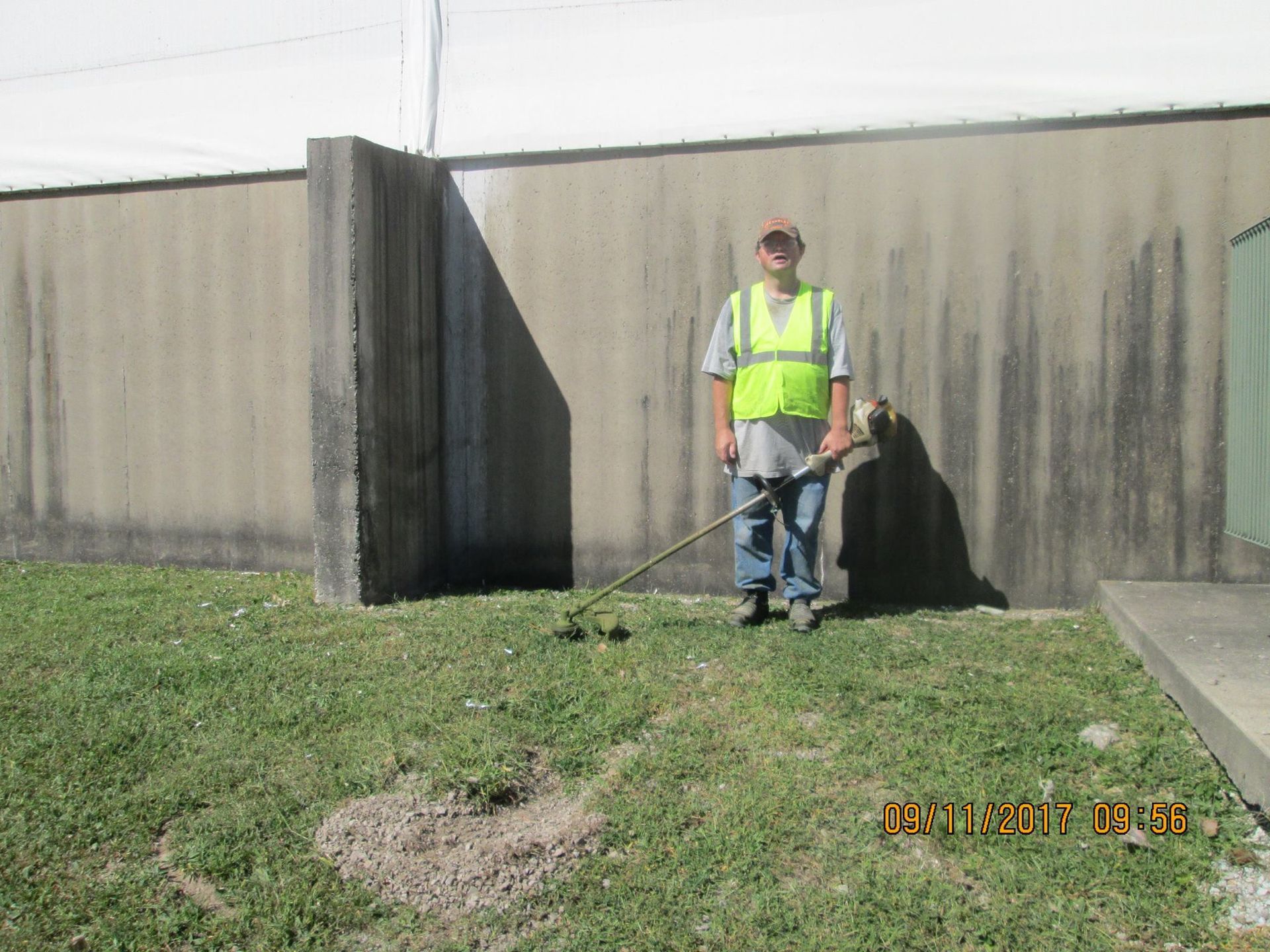 Person in safety vest trimming grass near a building with a weed whacker.