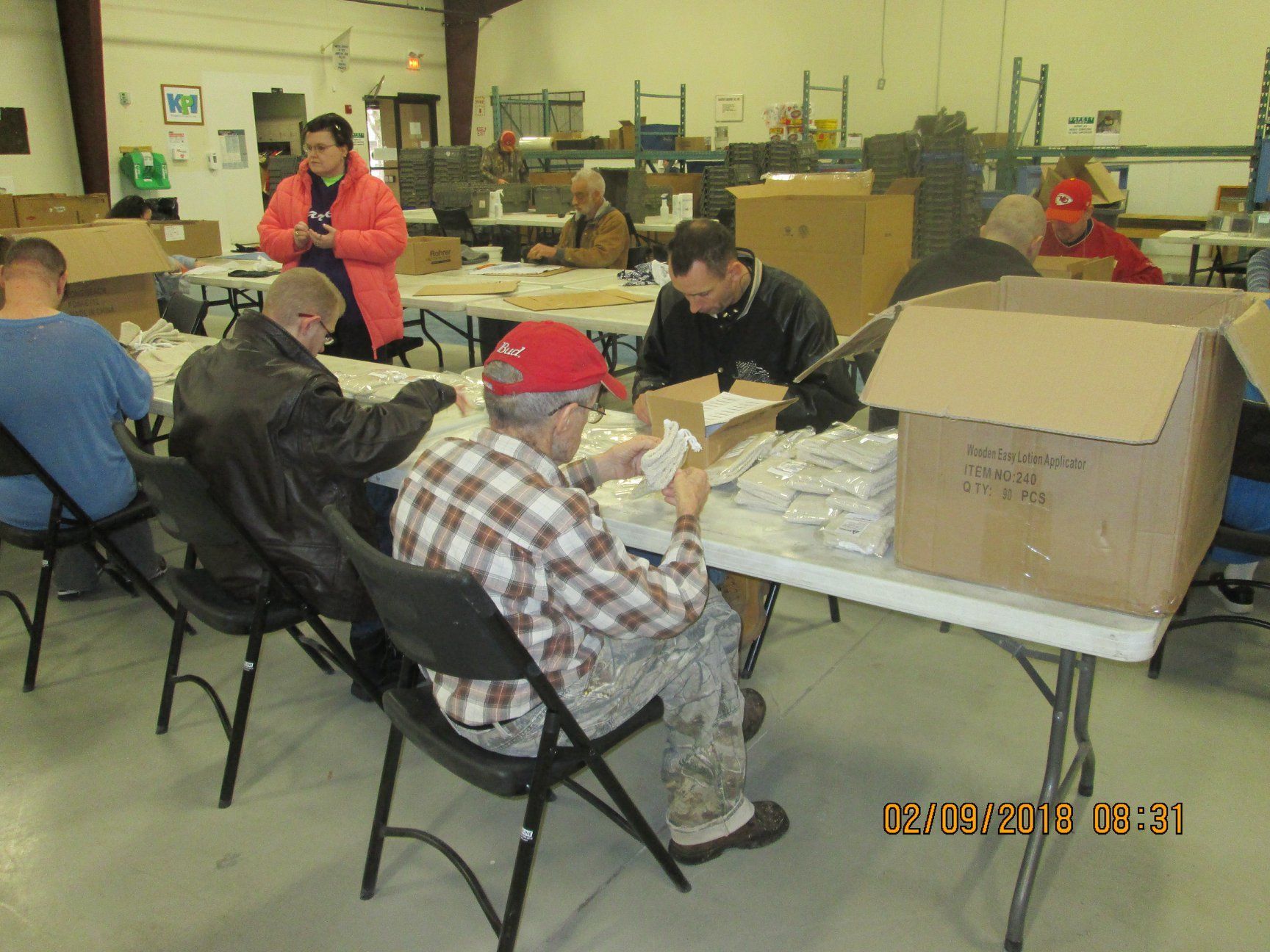 People packing items at tables in a warehouse setting. Boxes and supplies are present. People packing items at tables in a warehouse setting. Boxes and supplies are present.