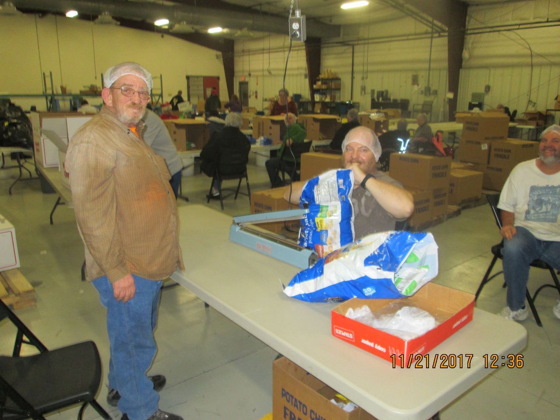 People in hairnets packing food items at tables in a warehouse.