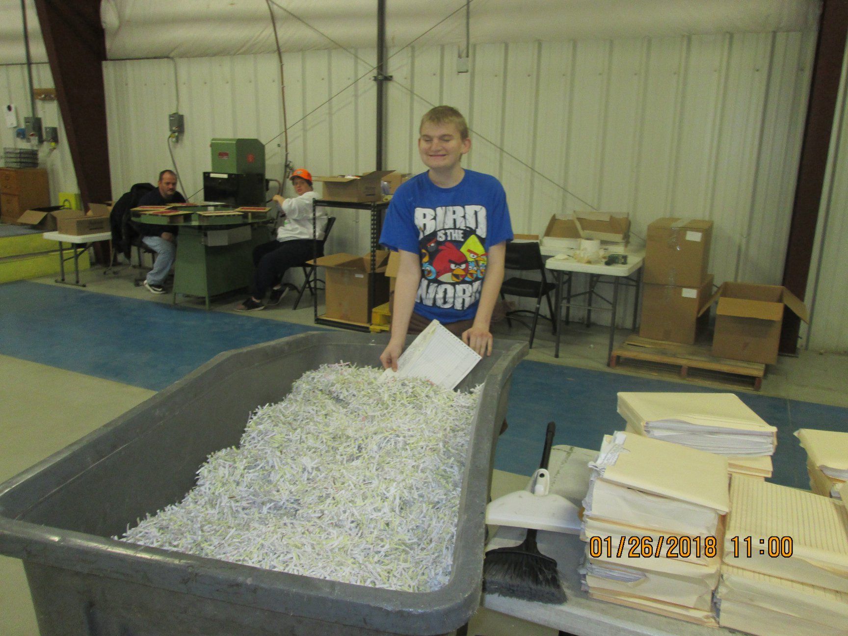 Boy with shredded paper, stands by a bin in a workshop. Others work at tables. Boy with shredded paper, stands by a bin in a workshop. Others work at tables.