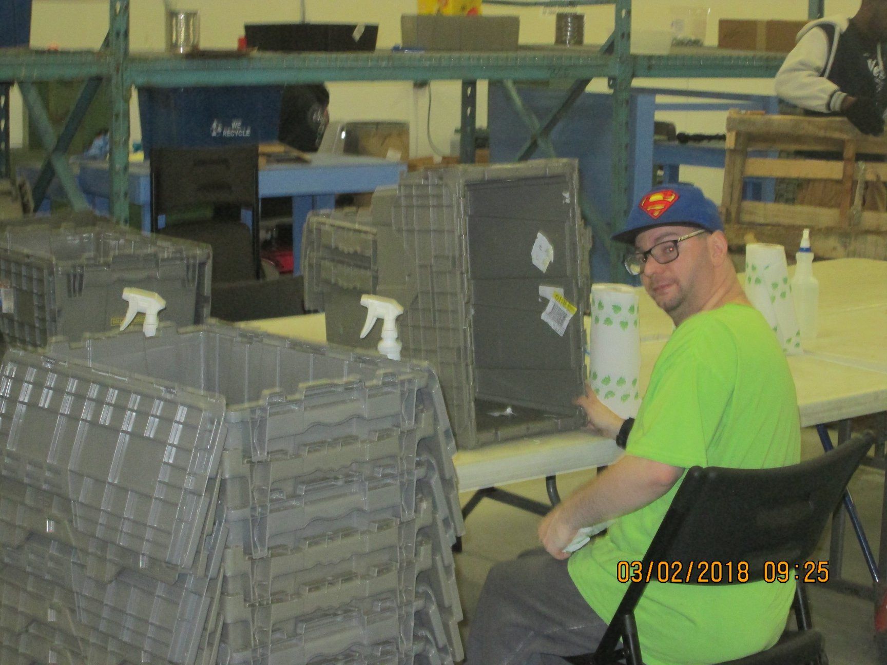 Man in green shirt, Superman hat, working at a table in a warehouse, crates stacked nearby.