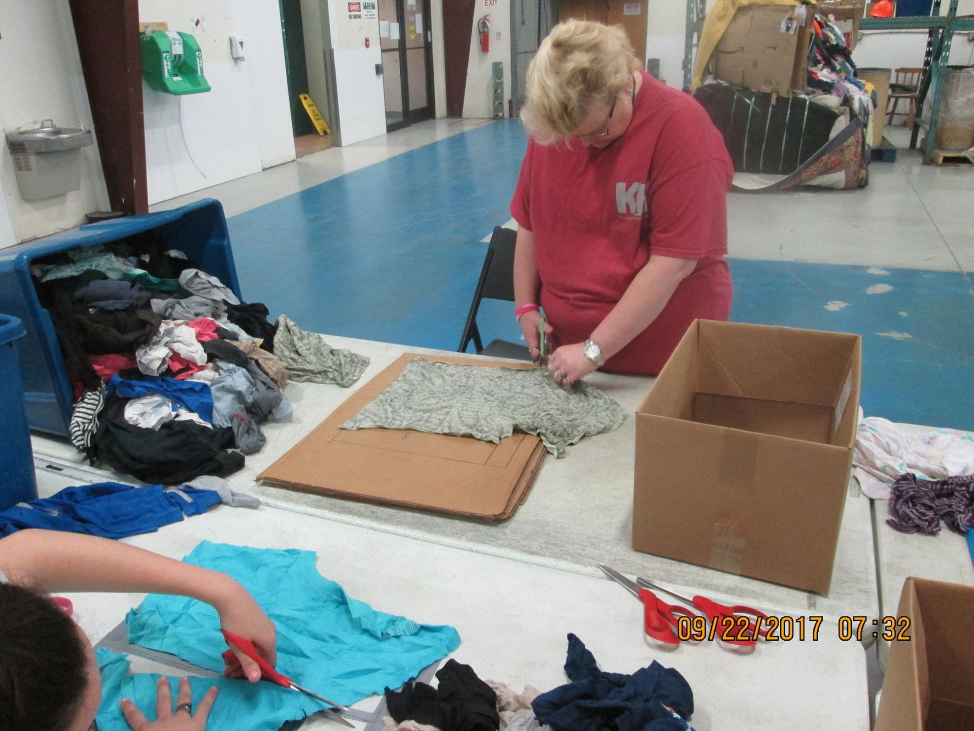 People cutting clothing at a table, likely for donation or repurposing. Blue floor, cardboard boxes, various fabrics.
