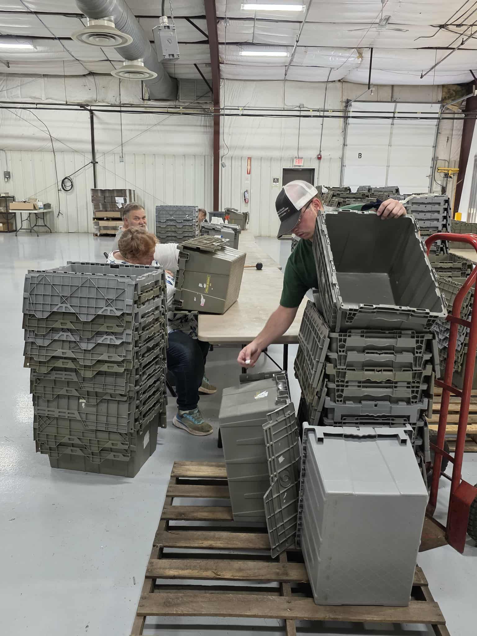 Two people stack gray plastic containers in a warehouse. A man in a cap lifts one; another person sits at a table. Two people stack gray plastic containers in a warehouse. A man in a cap lifts one; another person sits at a table.