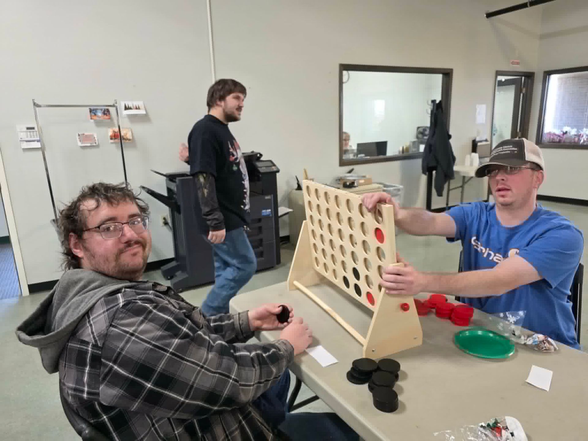 Three people playing a game of Connect Four at a table in a room. One man is looking at the camera.