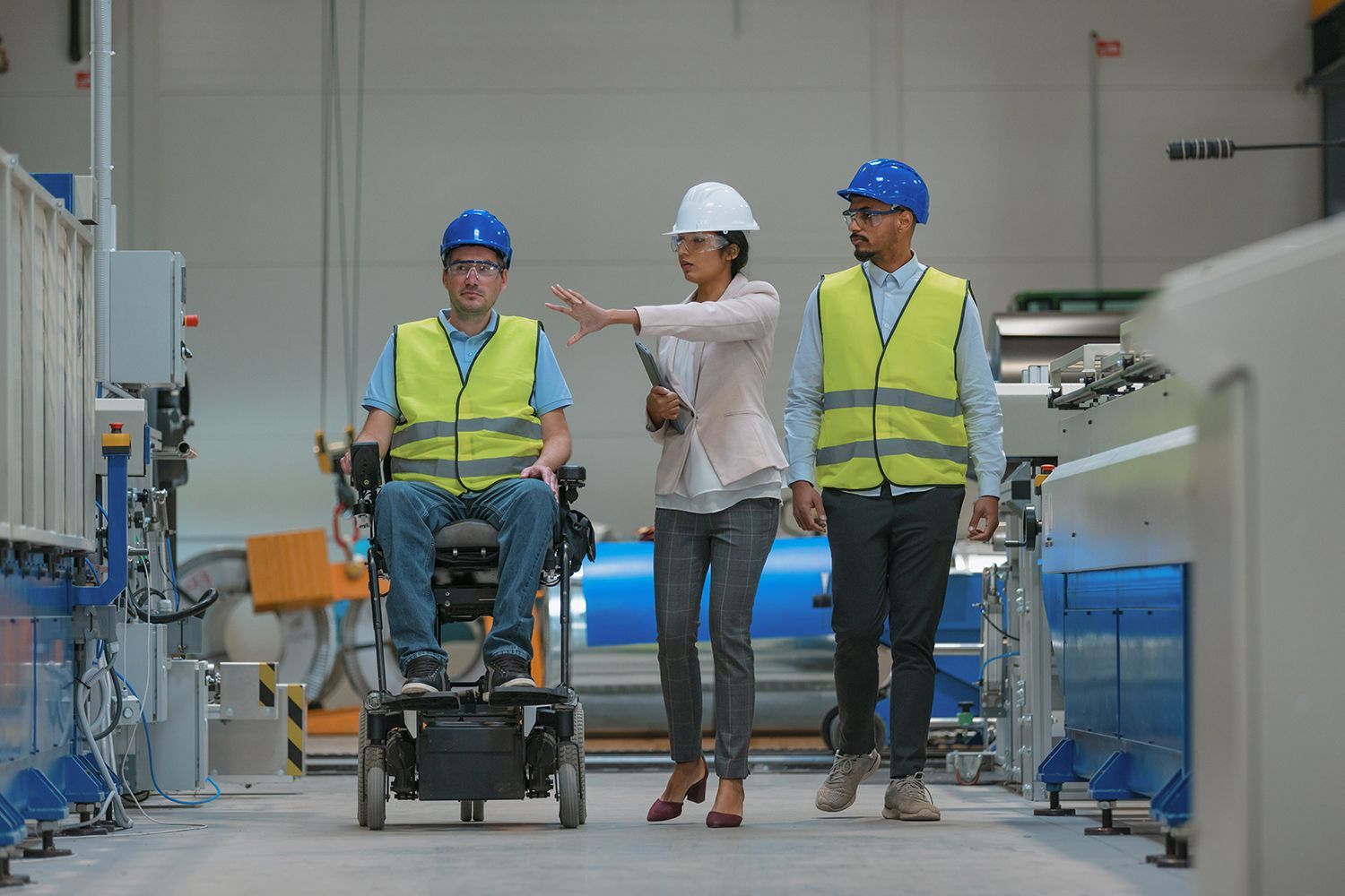 A factory floor with three people: one in a wheelchair, two walking, all wearing hard hats and safety vests. A factory floor with three people: one in a wheelchair, two walking, all wearing hard hats and safety vests.