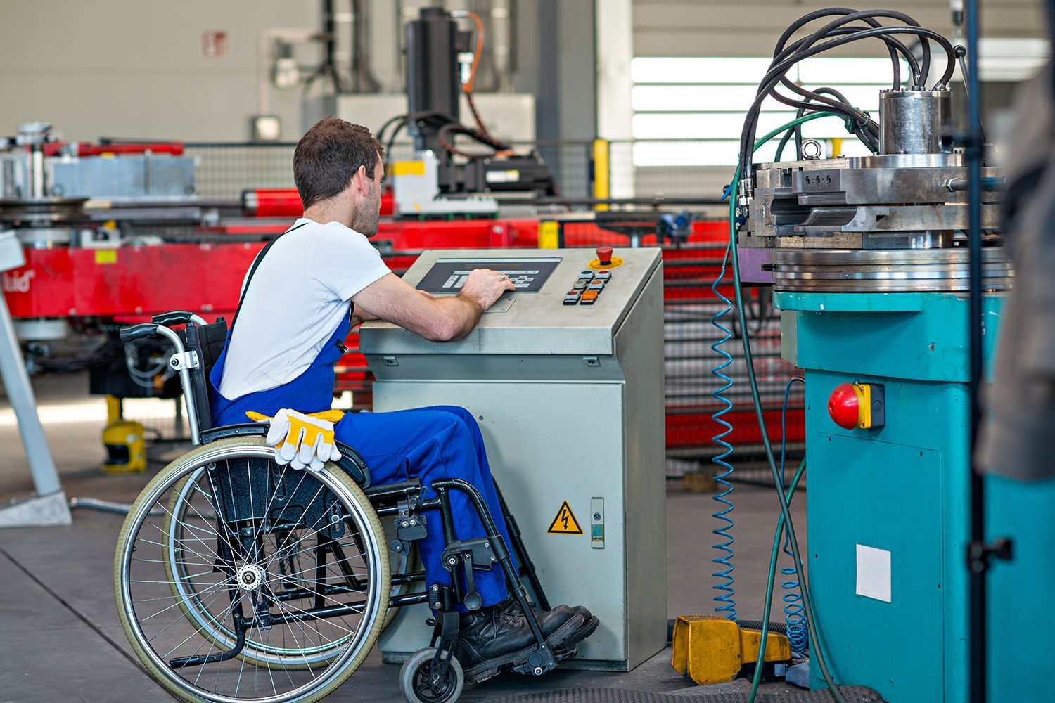 Man in wheelchair operating machinery in a factory; control panel, equipment, and wires visible.