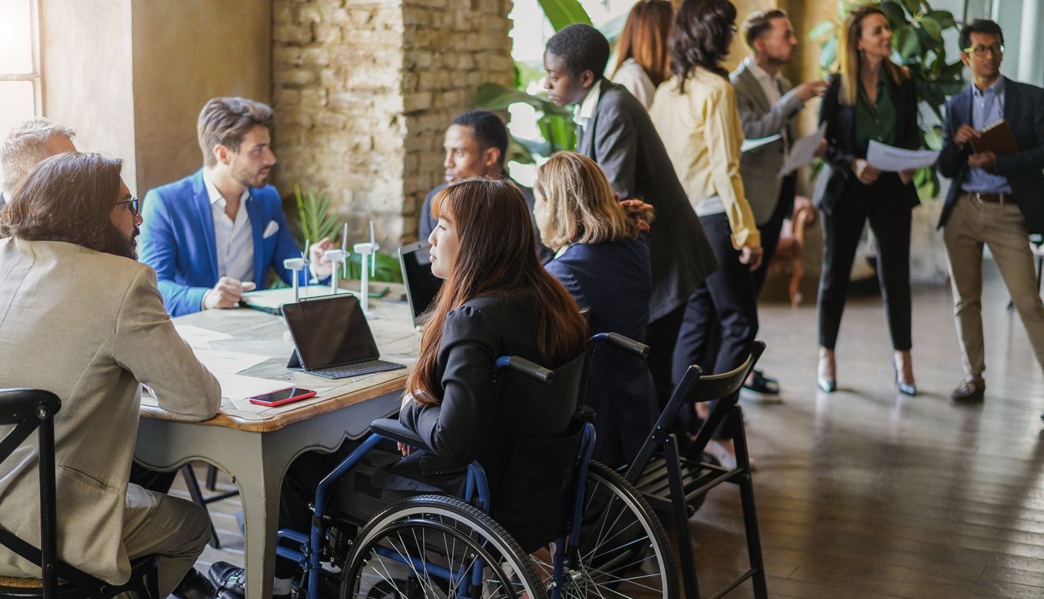 Business meeting: People at a table, one in a wheelchair, others standing and discussing in a modern office. Business meeting: People at a table, one in a wheelchair, others standing and discussing in a modern office.