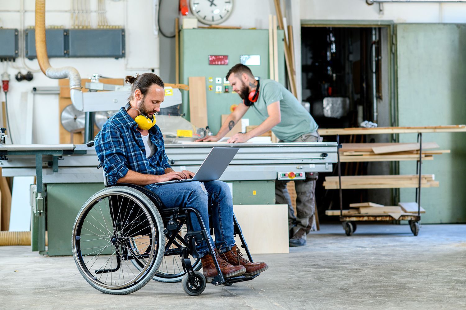 Man in wheelchair using a laptop in a woodshop while another man works at a table saw. Man in wheelchair using a laptop in a woodshop while another man works at a table saw.