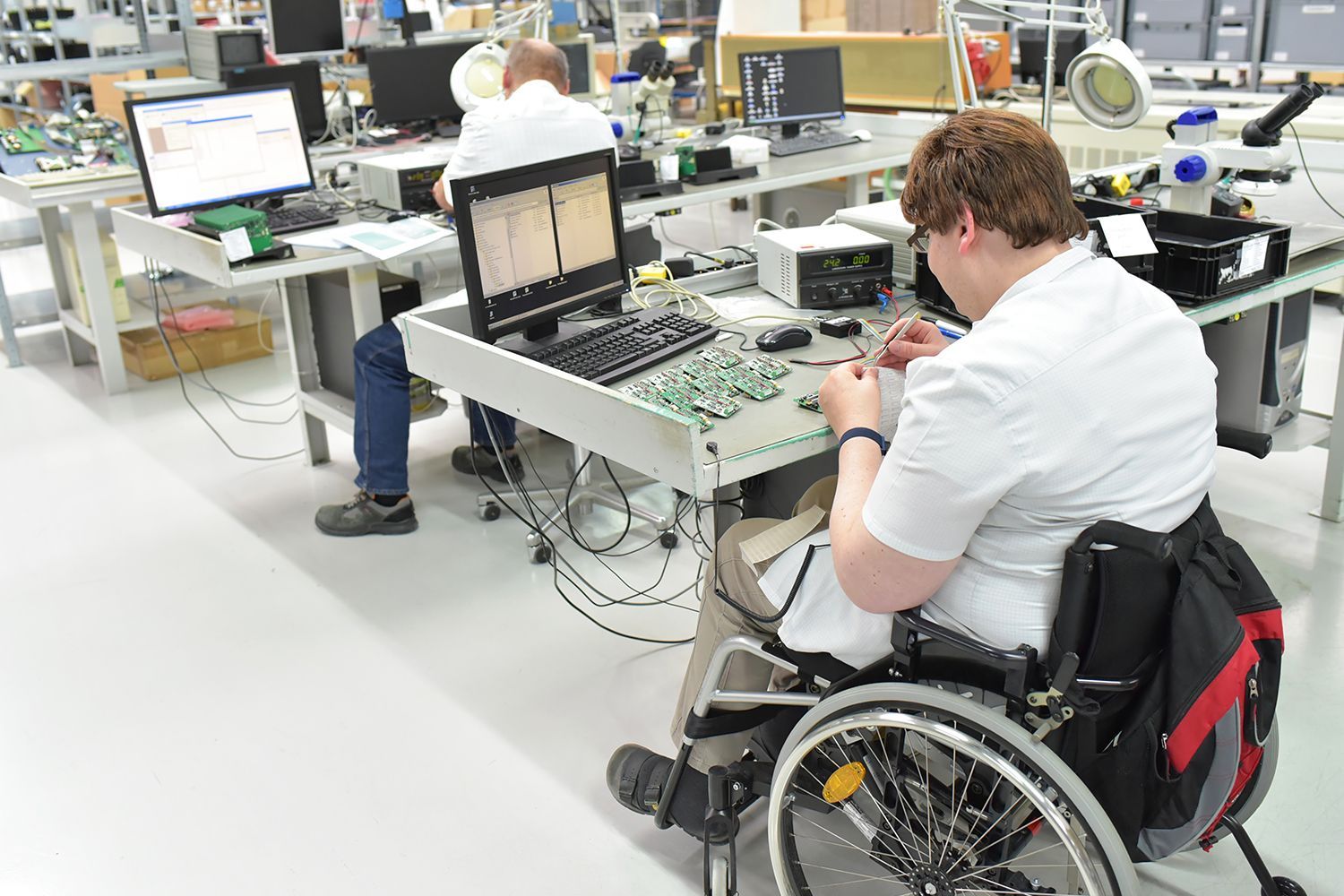 Woman in wheelchair working with electronics at a workstation. Another person works in the background. Factory setting. Woman in wheelchair working with electronics at a workstation. Another person works in the background. Factory setting.