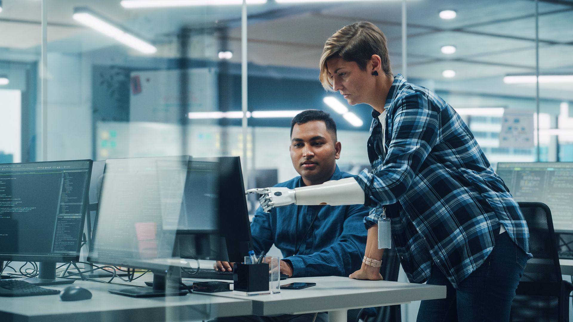Two people at computers, one with a robotic arm, working in an office. Two people at computers, one with a robotic arm, working in an office.