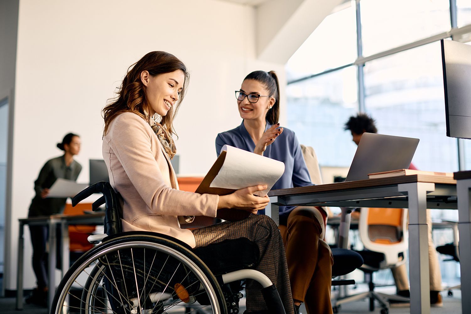 Woman in wheelchair and colleague review documents in office. Woman in wheelchair and colleague review documents in office.