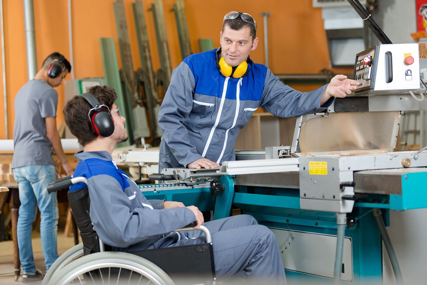 Man in wheelchair operates saw, instructed by a man in a workshop. A second man watches. All wear hearing protection. Man in wheelchair operates saw, instructed by a man in a workshop. A second man watches. All wear hearing protection.
