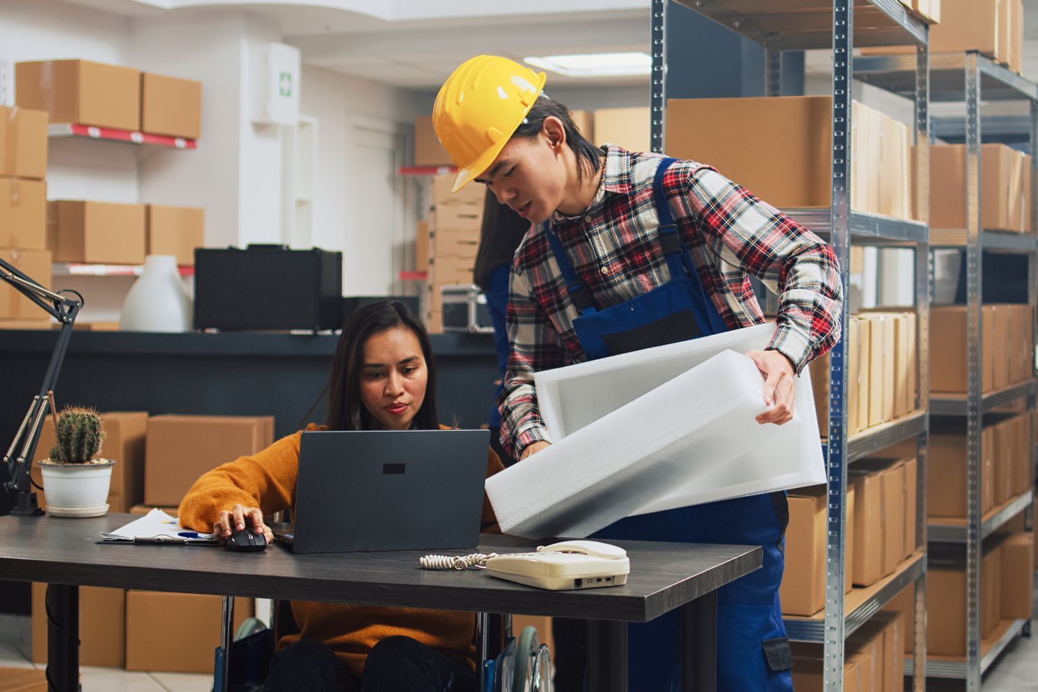 Woman in wheelchair uses laptop while worker in hard hat holds a white container in a warehouse setting. Woman in wheelchair uses laptop while worker in hard hat holds a white container in a warehouse setting.
