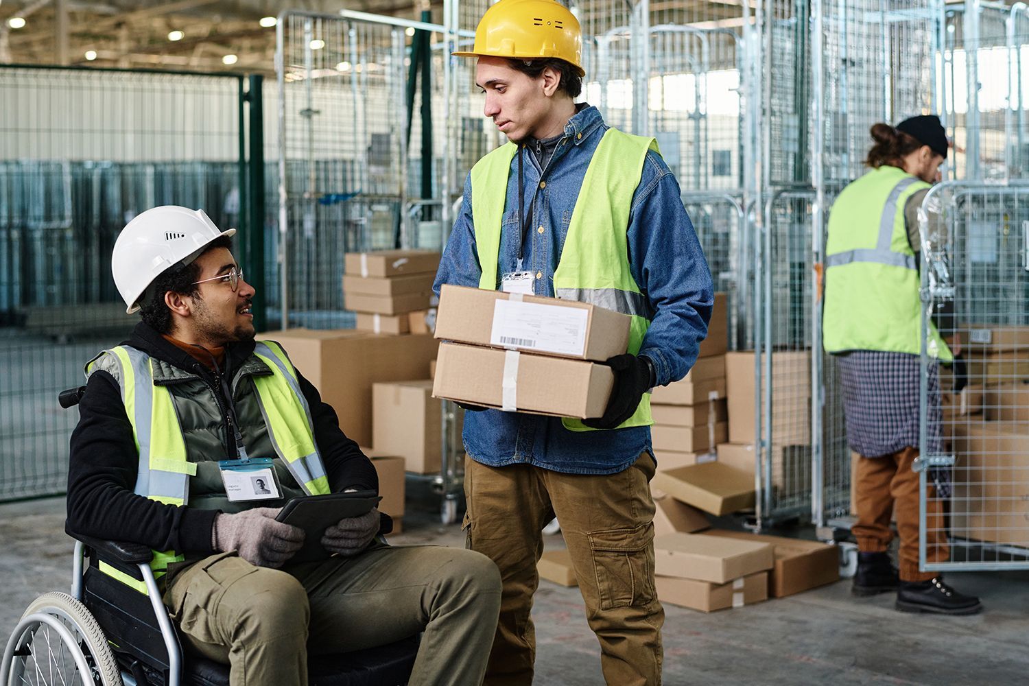 Warehouse workers: one in a wheelchair, talking to another carrying packages, a third near a rolling cart. Warehouse workers: one in a wheelchair, talking to another carrying packages, a third near a rolling cart.