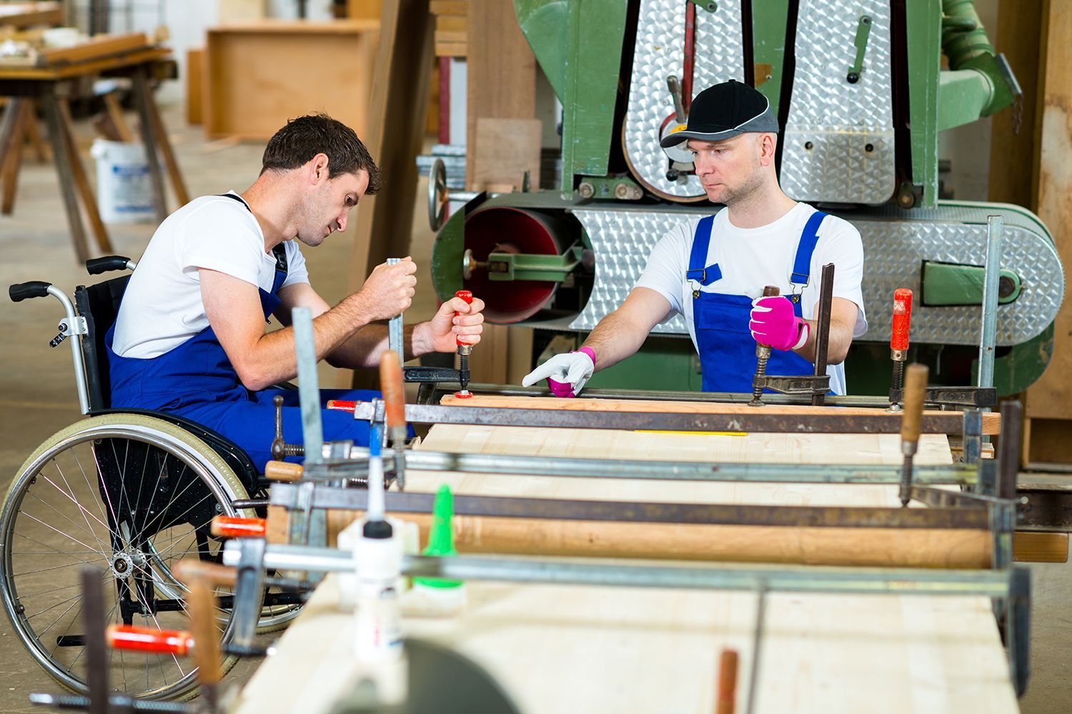 Two men working in a wood shop; one in a wheelchair, using tools at a workbench near machinery.