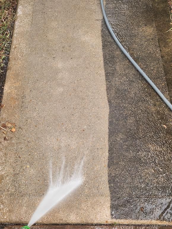 Person pressure-washing a concrete walkway, cleaning a dark stained section beside a hose.