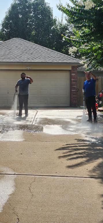 Two people standing beside a wet driveway and garage, with water spraying across the pavement.
